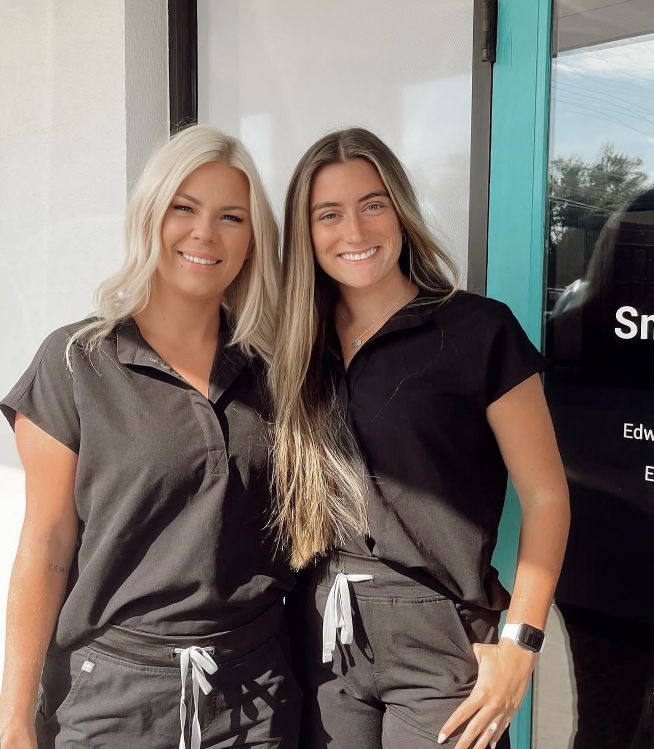 Two women standing side by side in front of a storefront, smiling at the camera both are wearing black shirts with white emblems on their chests and black pants.