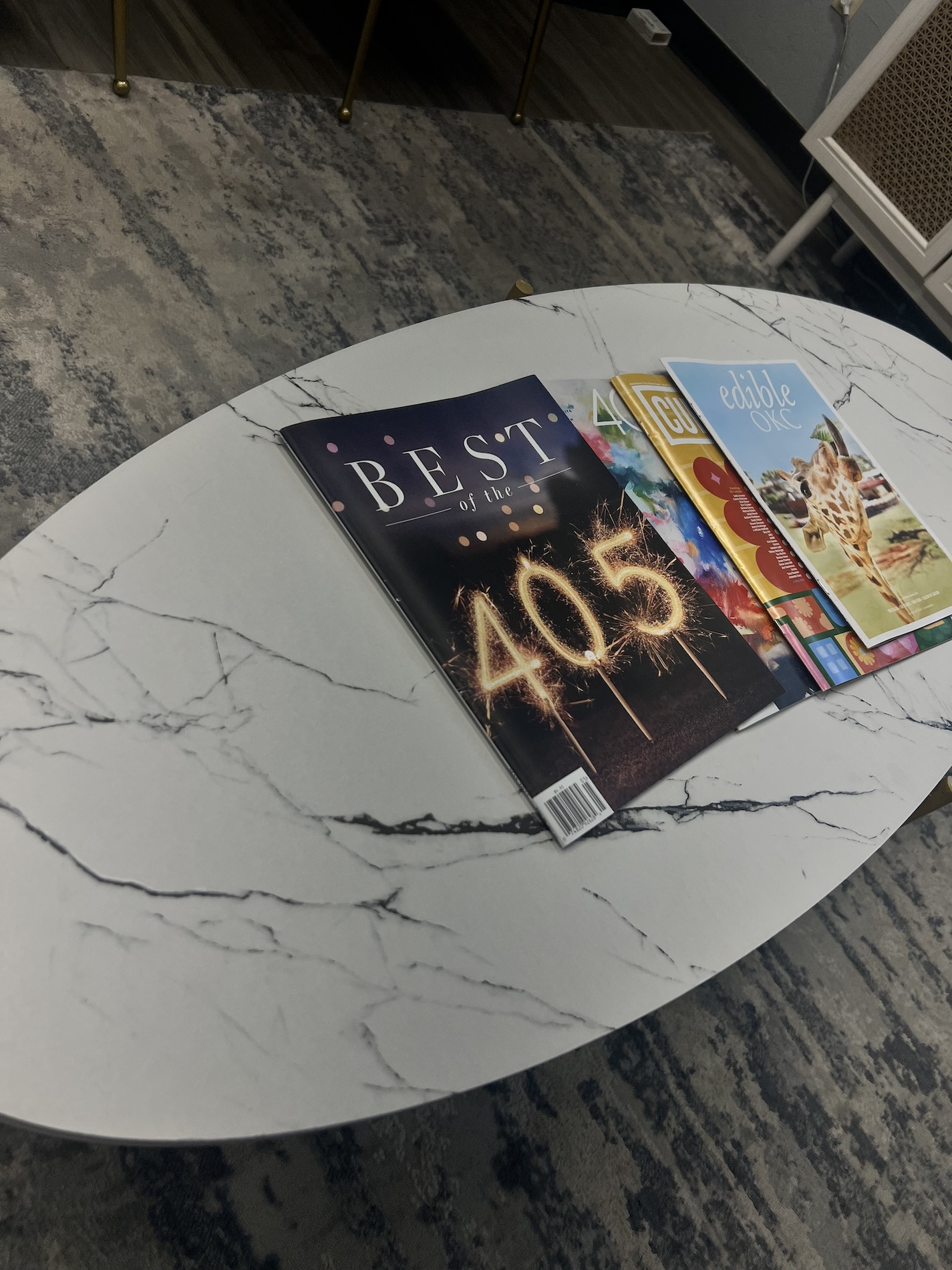 The image shows a coffee table with various items on top, including magazines, books, and a framed certificate, all arranged neatly on a marble surface.