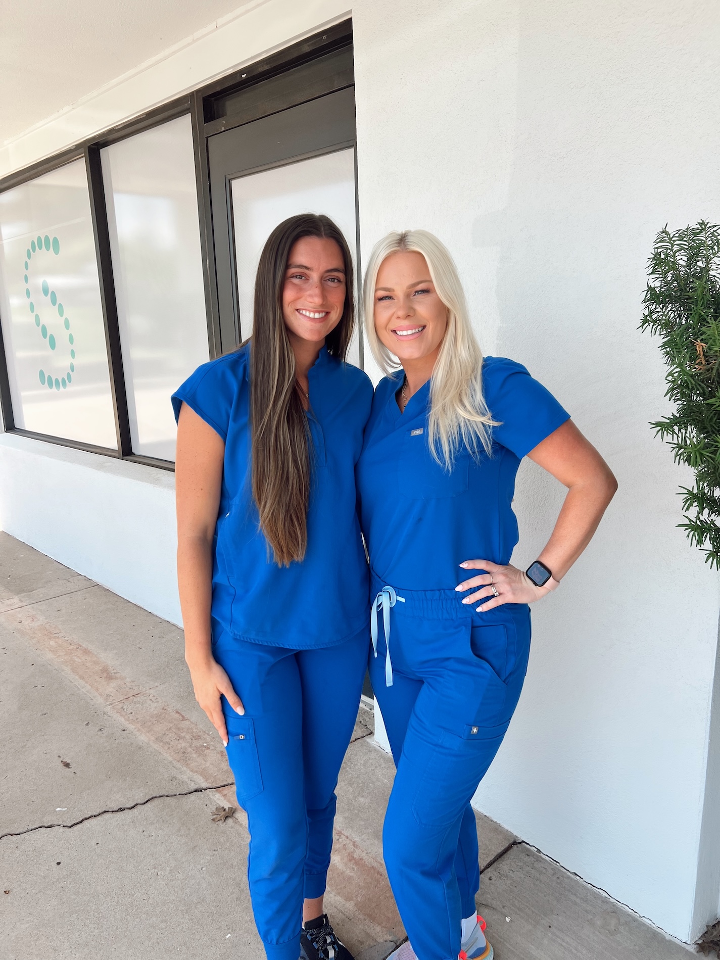 Two women wearing scrubs stand together smiling, posing for a photo outside a building with a white wall and green trim.