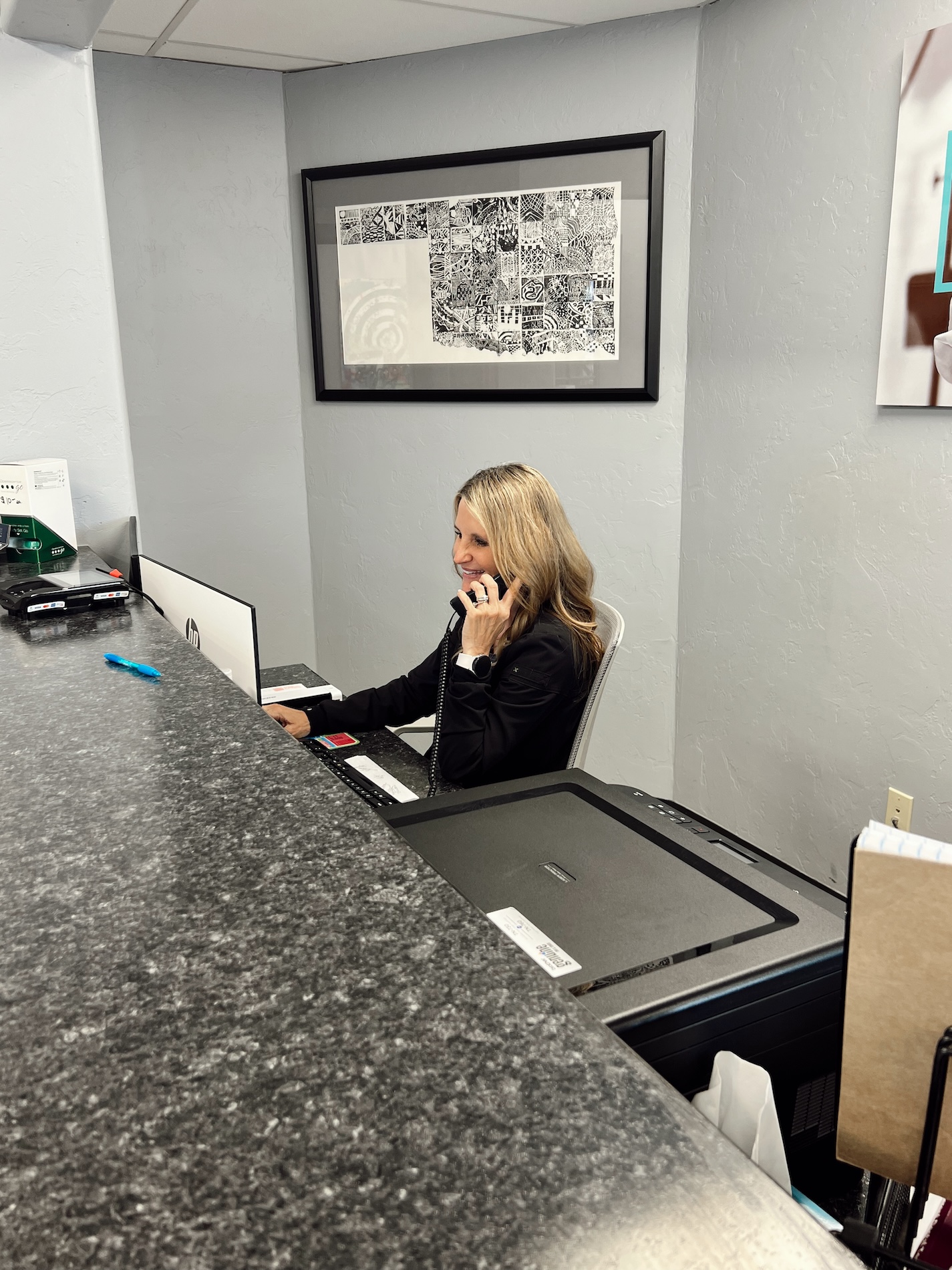 A woman is sitting at a desk in an office environment, using a computer with a keyboard and mouse, while talking on her cell phone.