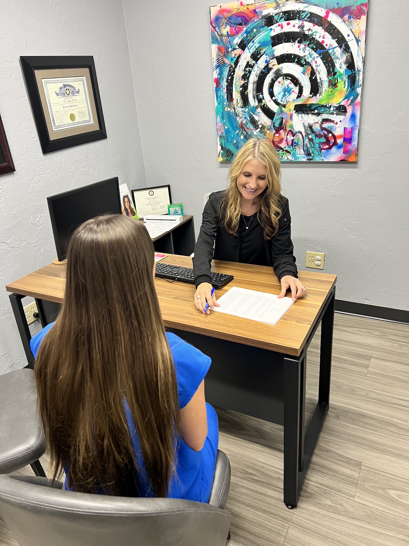 Woman sitting at desk with papers, signing document while smiling at girl standing in front of her.