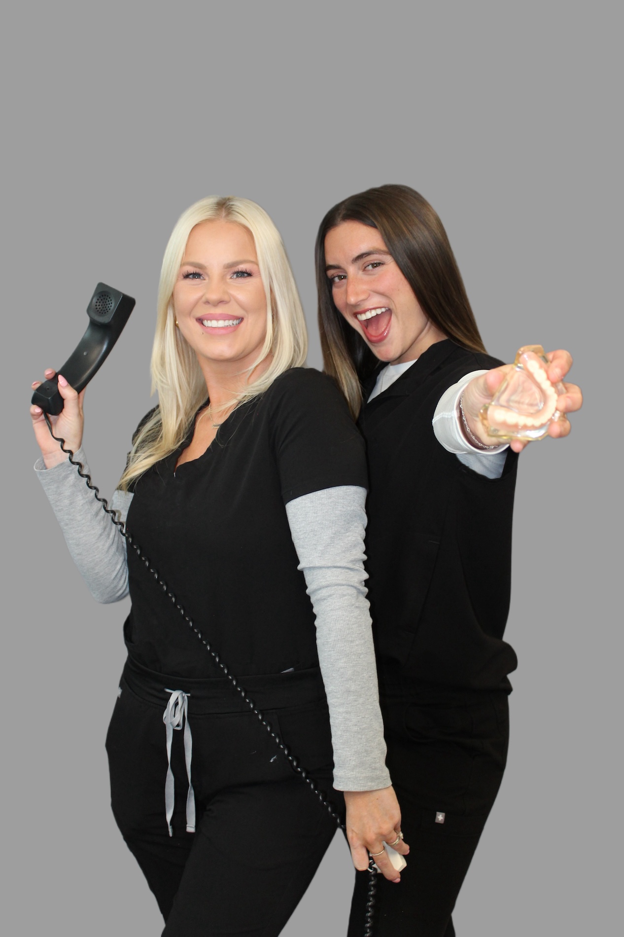 Two women wearing scrubs, one holding a phone, smiling at the camera.