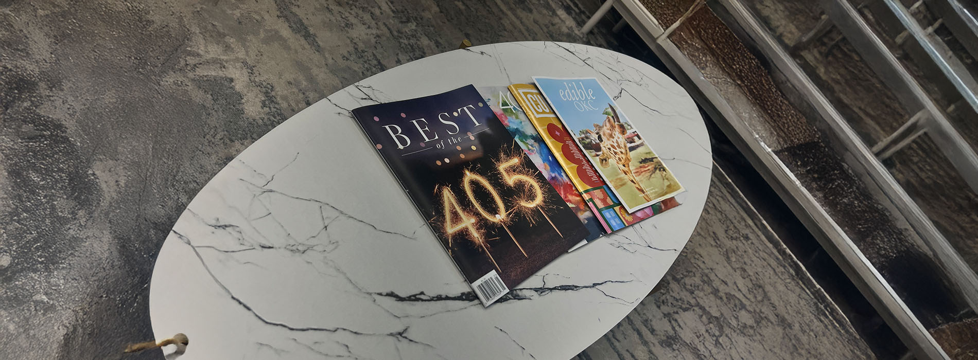 The image features a table with an assortment of books and magazines displayed on it, placed against a background that appears to be a wall with a marble-like texture.