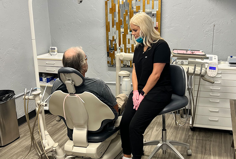 A dental office with a dentist seated at a chair and a patient standing beside it, both wearing face masks.