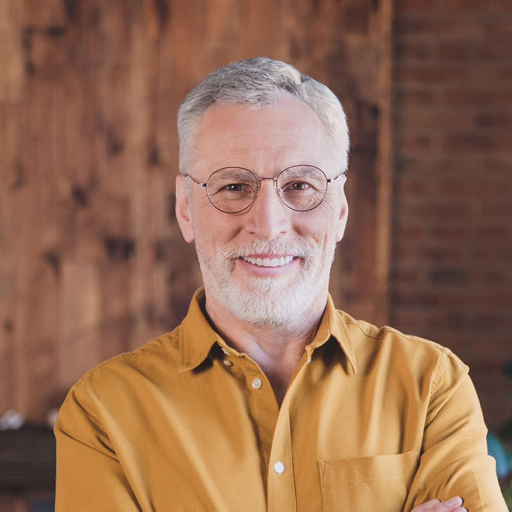 The image features a man with gray hair and a beard, wearing glasses and a yellow shirt, standing confidently with his arms crossed.