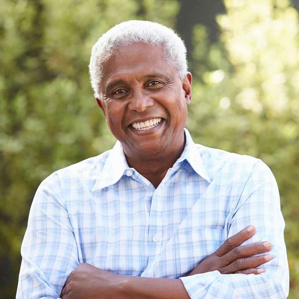 The image shows a smiling man with grey hair, wearing a light blue shirt, standing outdoors with his arms crossed.