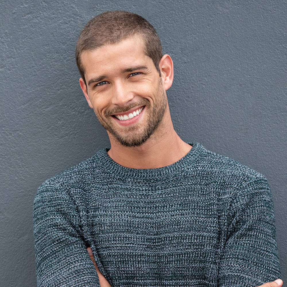 The image features a man with short hair and a beard, smiling at the camera, wearing a dark blue sweater over a lighter top, standing against a gray wall.