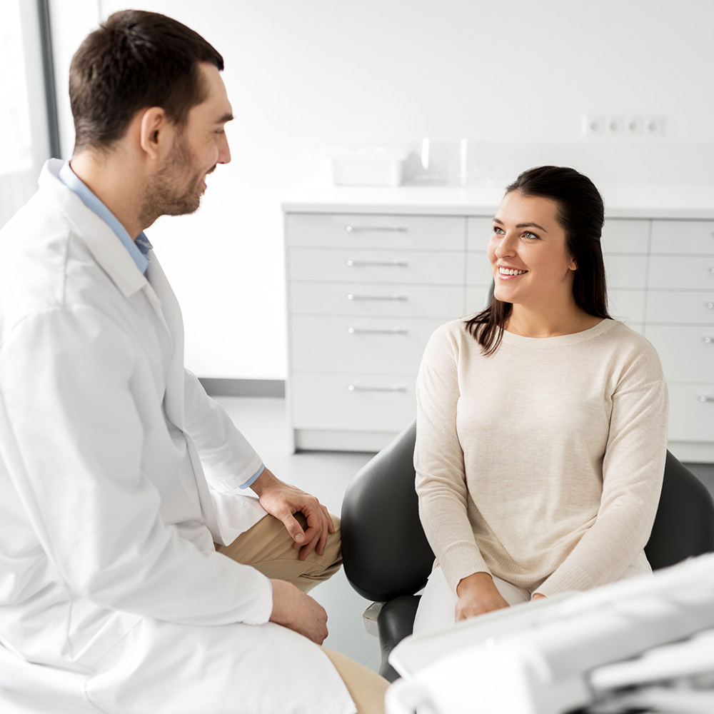The image shows a professional setting with a man standing behind a woman who is seated on a dental chair, both engaged in conversation  the man appears to be a dentist given his attire, which includes a white coat and stethoscope, while the woman is dressed casually in a long-sleeved shirt.