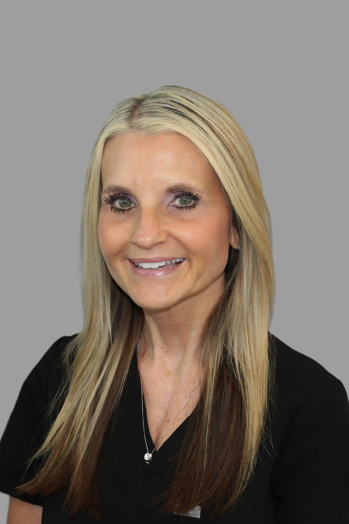 A woman with long blonde hair wearing a white shirt and a necklace, smiling at the camera.