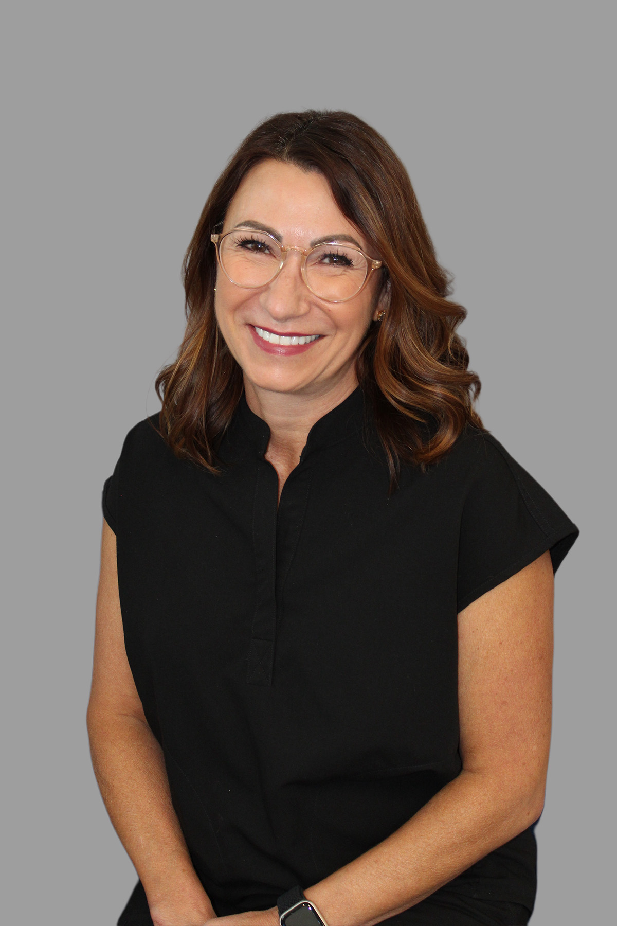 A woman with glasses is sitting, smiling slightly, with her hands on her lap, wearing a black shirt, against a neutral background.