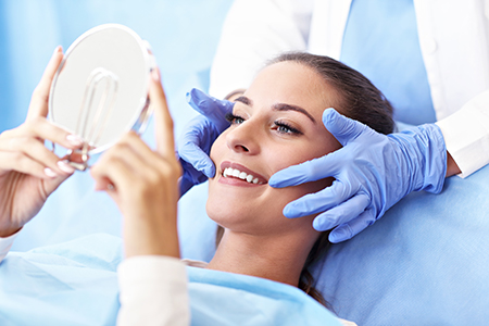 A woman receiving a facial treatment with a magnifying mirror held by two professionals, one standing and one sitting at her side.